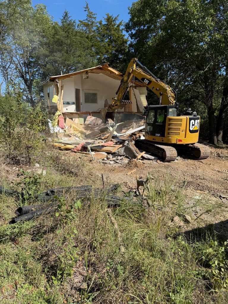 Yellow excavator demolishing a small white house in a wooded area. Debris and exposed structure visible.
