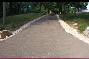 Asphalt driveway lined with white gravel, leading toward trees and a house on a sunny day.
