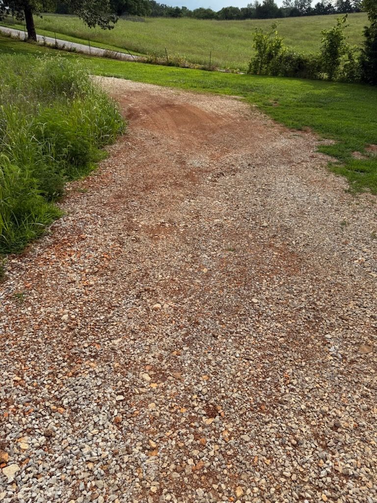 Gravel driveway leading into a grassy field. Reddish-brown and grey stones, green grass, trees and sky.