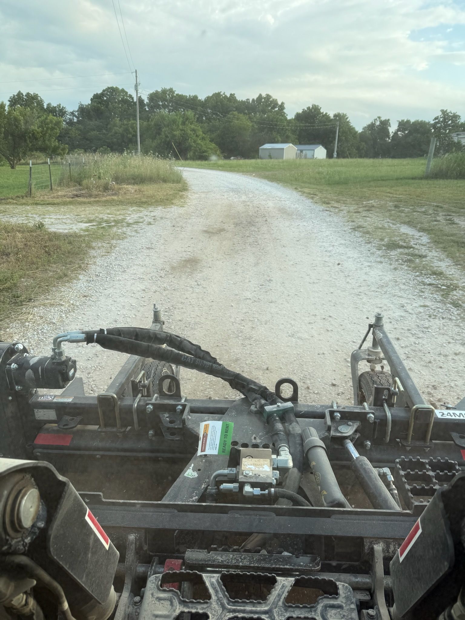 A tractor grading a gravel road on a cloudy day, trees and a house in the background.