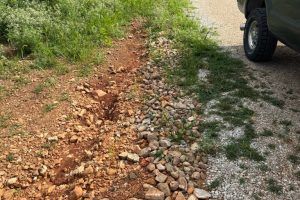 Ditch alongside a gravel road with loose rocks, dirt, and grassy vegetation; a vehicle tire is visible.