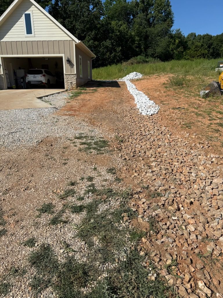 Gravel driveway leading to a garage. White rocks line a slope. Sunny day, beige garage.