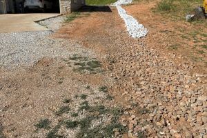 Gravel driveway with a rock-lined drainage channel on a hillside. The ground is reddish-brown.