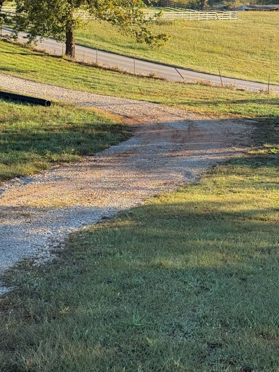 Gravel path leading through a grassy area, asphalt road and trees in the background, daytime.