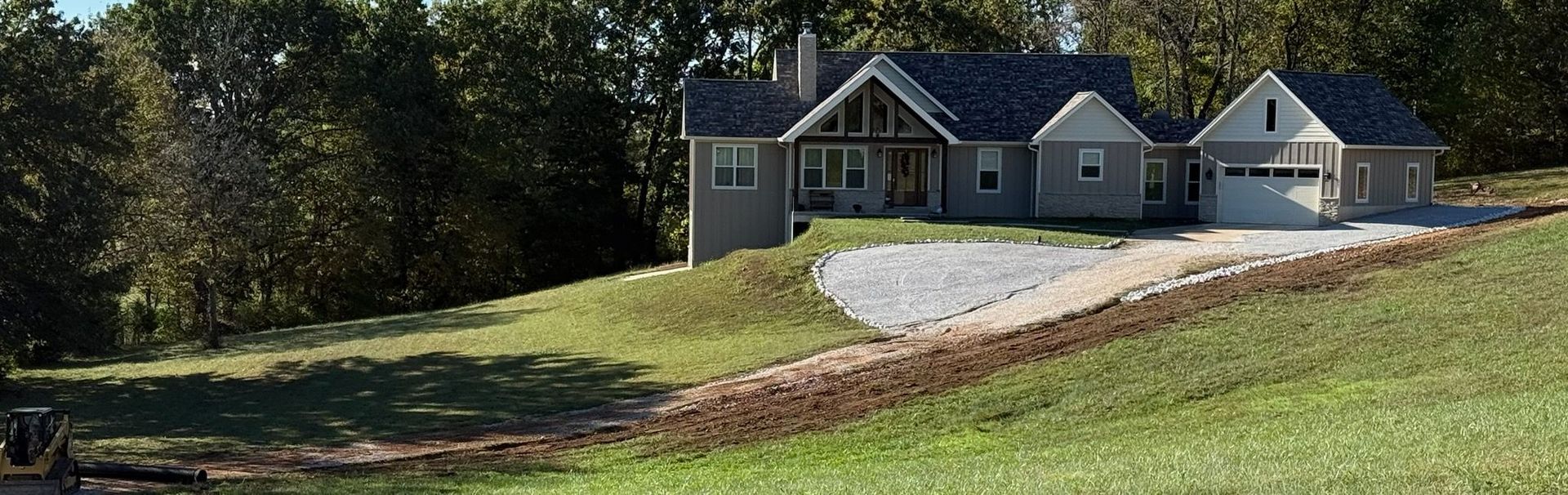House on a grassy hill with a gravel driveway. Trees in the background. Sunny day.