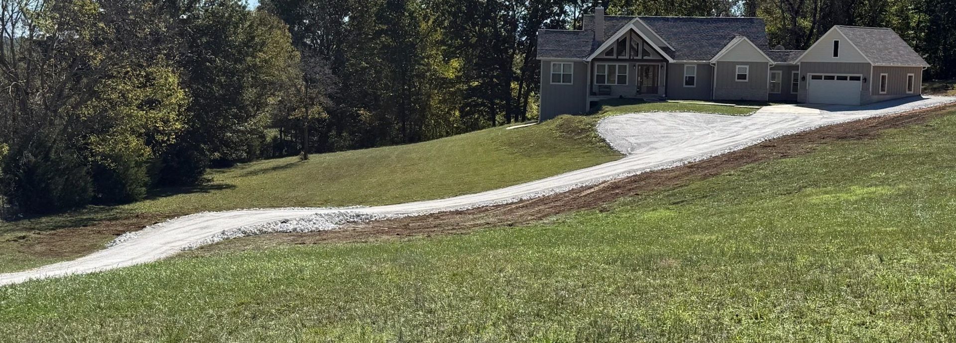 A house with a gravel driveway on a grassy hill, surrounded by trees.