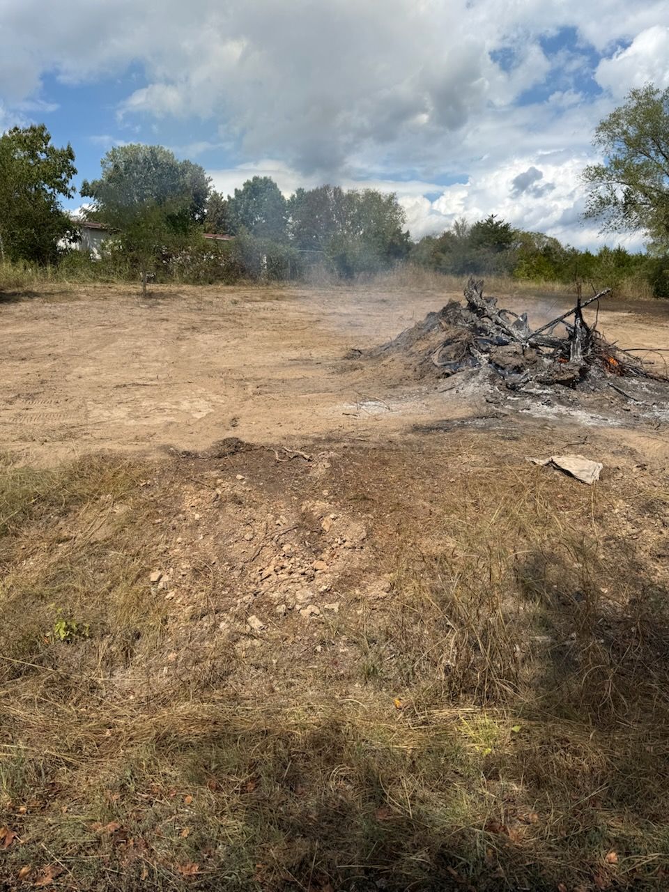 Smoldering debris pile in a cleared field, smoke rising, trees in background, overcast sky.