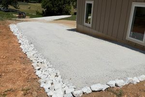 A gravel patio with a rock border next to a beige building with windows and an adjacent driveway.
