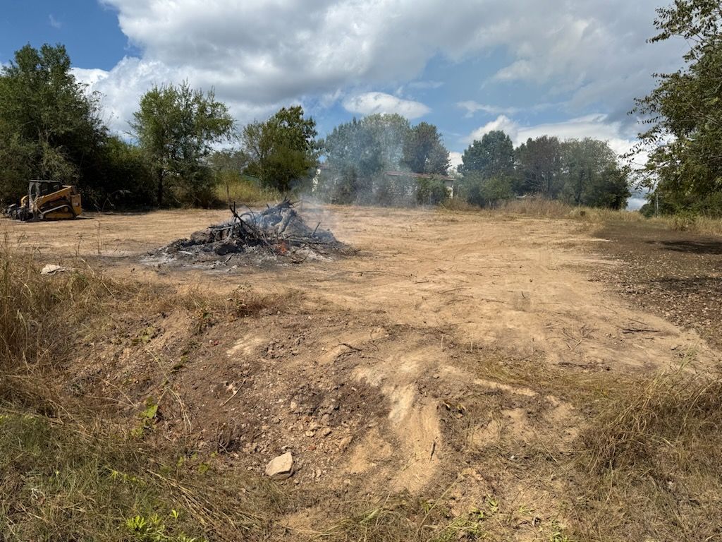 Pile of burning debris in a cleared, dry field. Trees in the background, blue sky with clouds overhead.