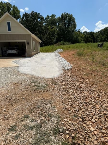 Gravel driveway leads to a beige house with a garage. Brown, grassy field in the foreground. Blue sky.