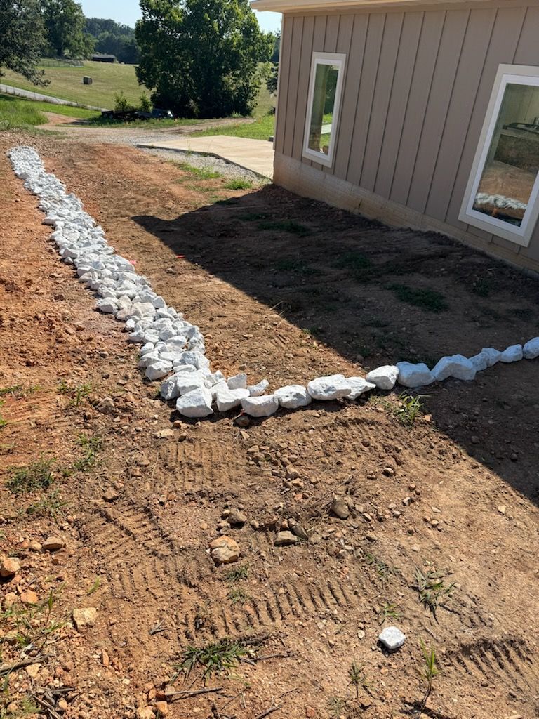 A dry creek bed made of white stones runs alongside a tan house on a grassy, dirt yard.