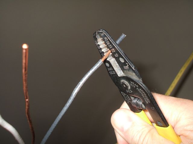 A hand using wire strippers to strip a black wire beside a bare copper wire.