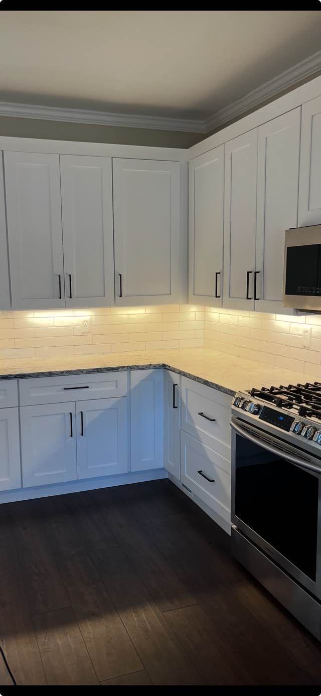 White kitchen with cabinets, dark wood floor, under-cabinet lighting, and a black stove in the corner
