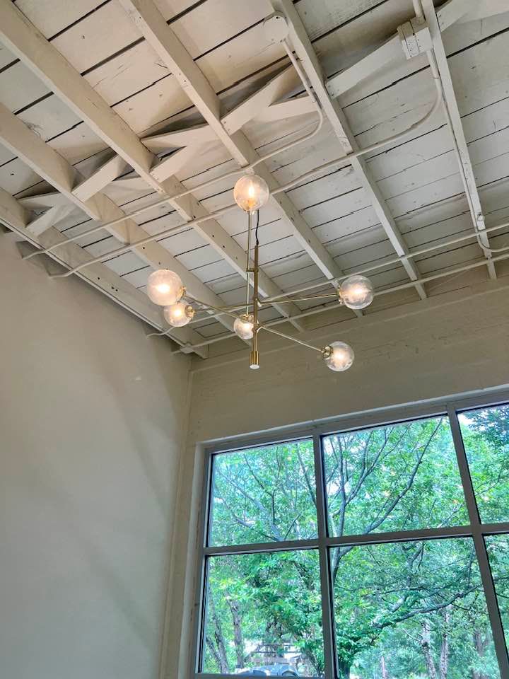 Modern ceiling light fixture in a bright room with white beams and large windows showing trees outside