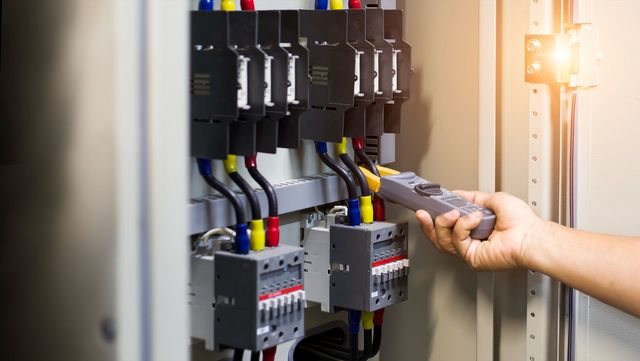Technician testing electrical control panel with a multimeter, wires and circuit breakers visible.