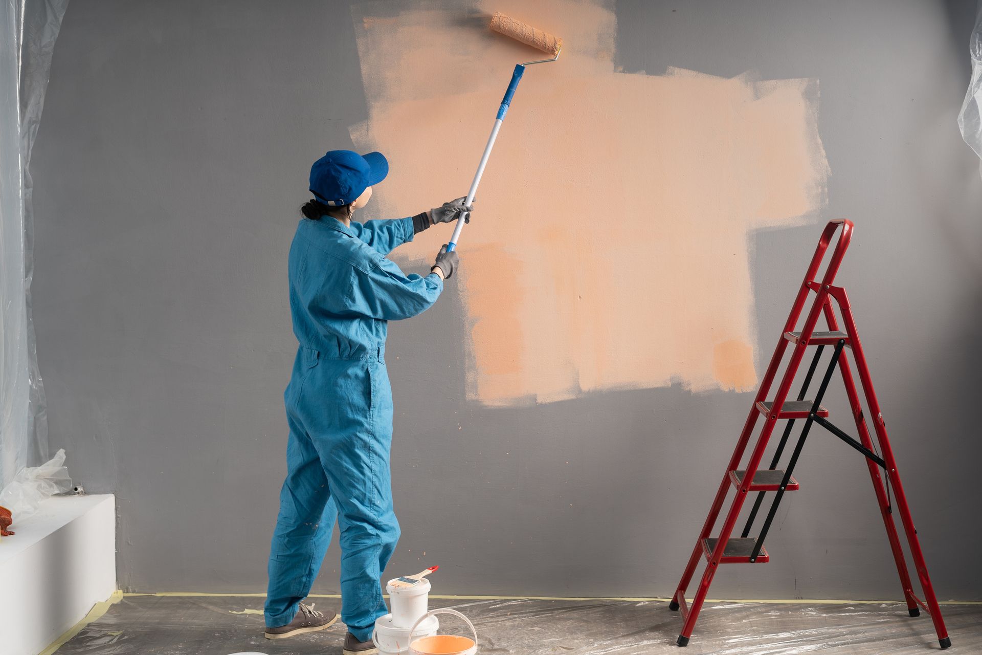 Man in blue shirt and white cap painting a wall light blue with a roller. Person in blue coveralls paints a wall with an orange roller, red ladder in the room.