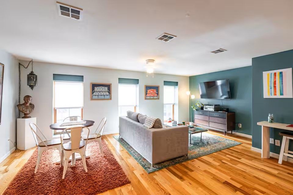 A living and dining area with hardwood floors, a gray sofa on a patterned rug, a dining table, and a teal accent wall.