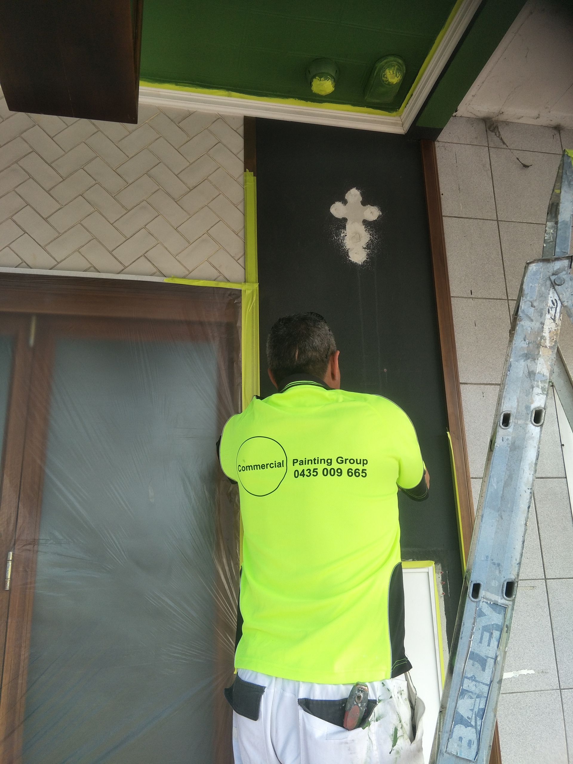 A man in a green shirt is on a ladder painting a wall — Commercial Painting Group in Thirroul, NSW