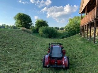 Red lawn tractor mowing grassy hill. Cloudy blue sky, trees, and wooden porch visible.