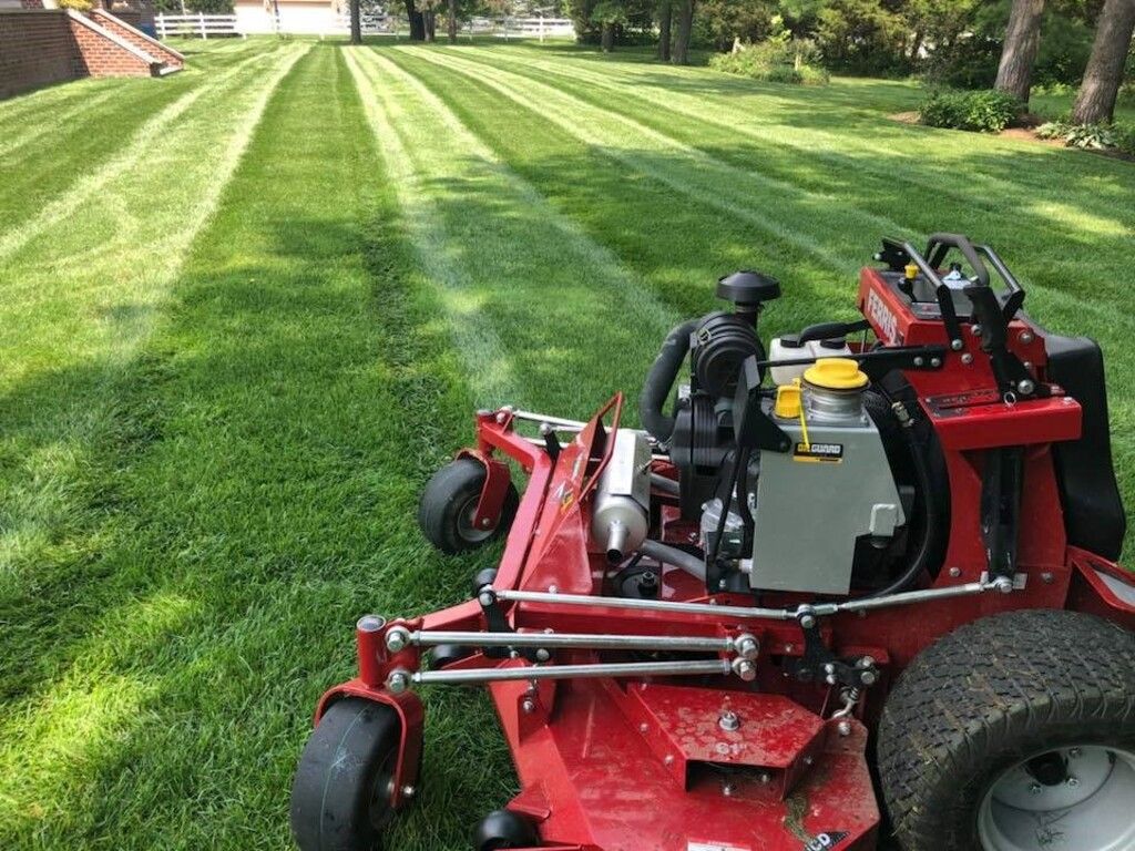 Red zero-turn mower stripes a green lawn; trees and a house are in the background.