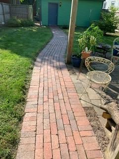 Brick pathway leads to a teal-colored shed, flanked by green grass and patio furniture.