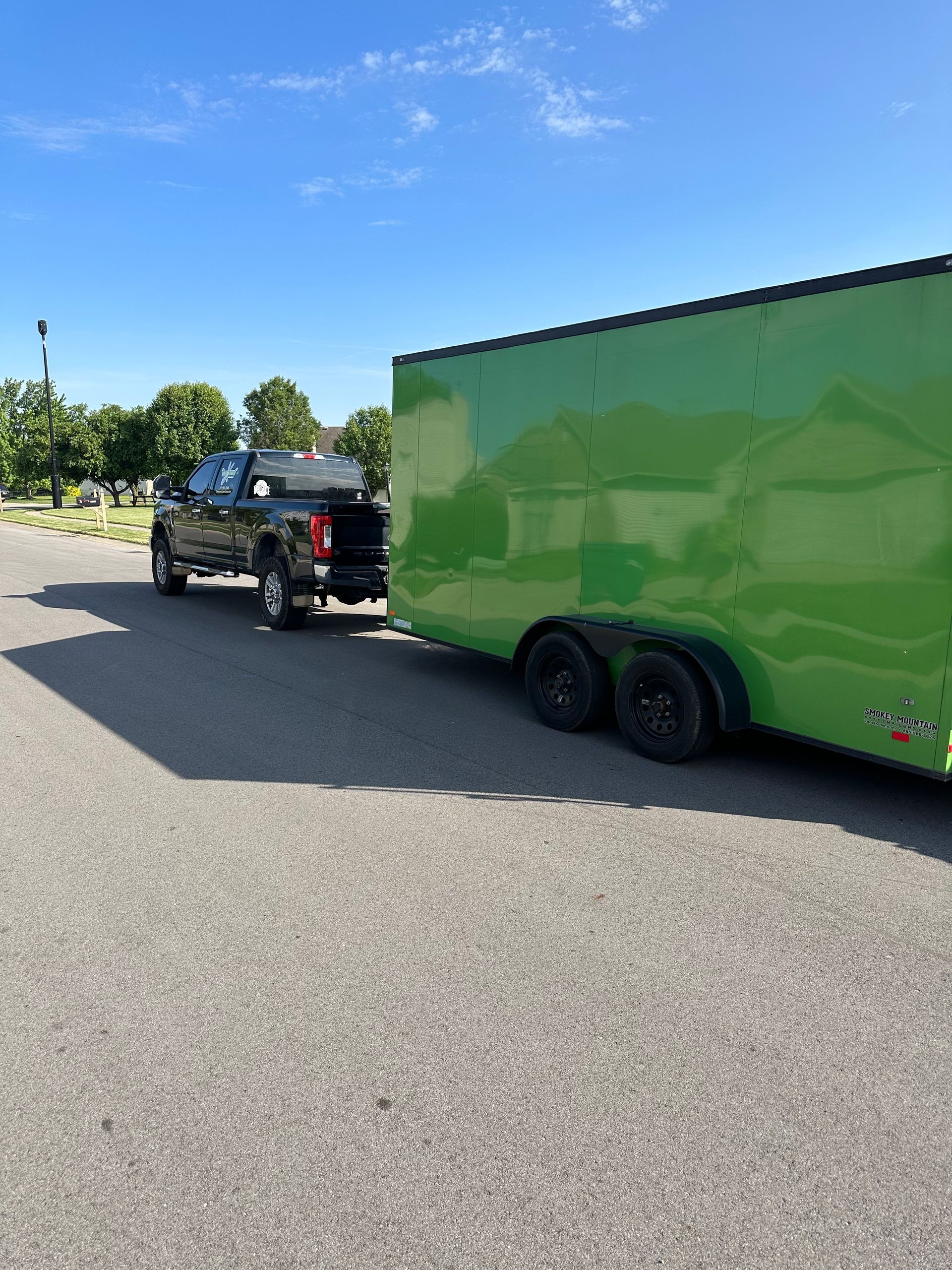 Dark truck towing a bright green enclosed trailer on an asphalt surface under a blue sky.