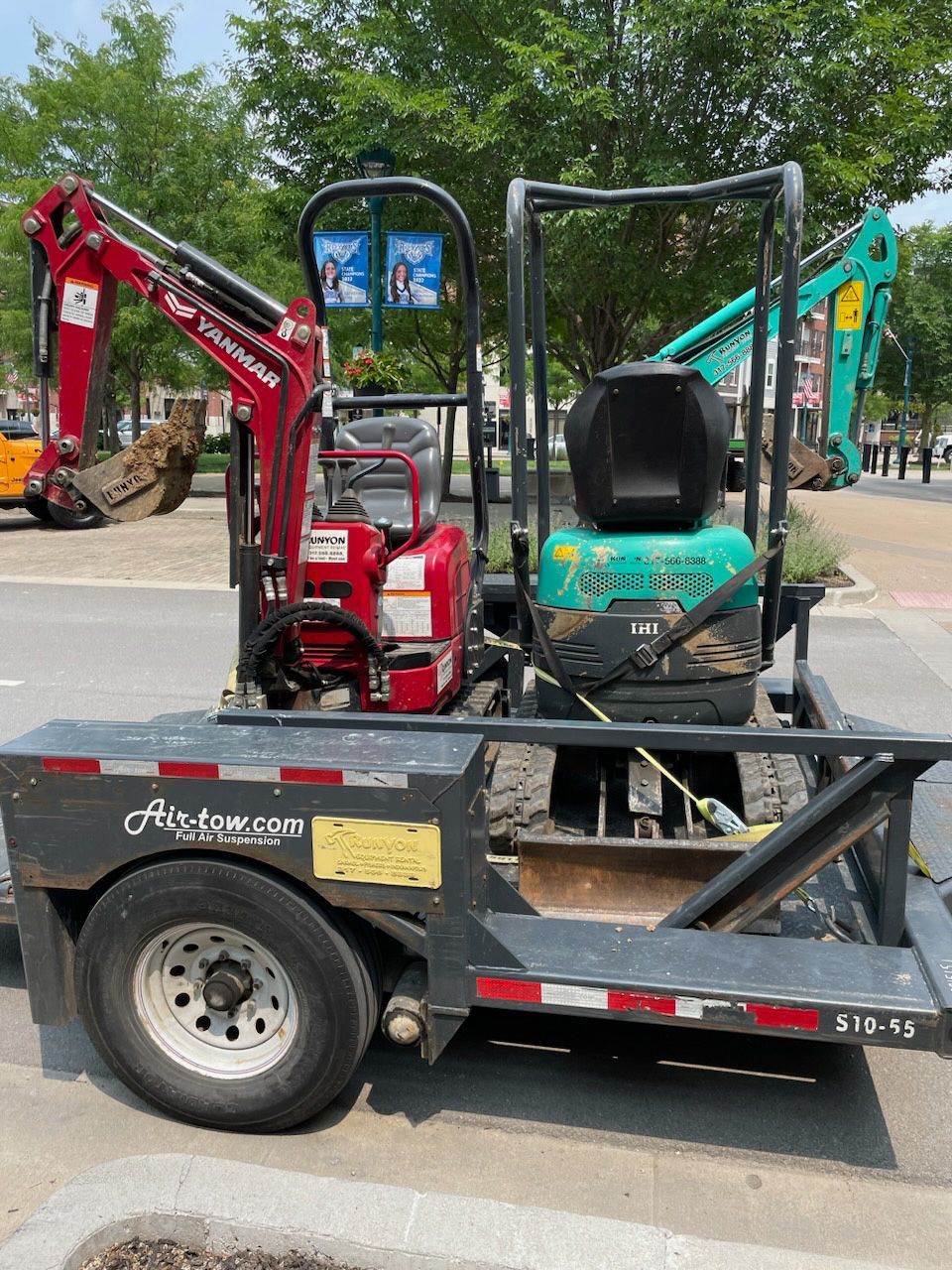 Two mini excavators on a trailer, red and teal, outdoors in a sunny environment.