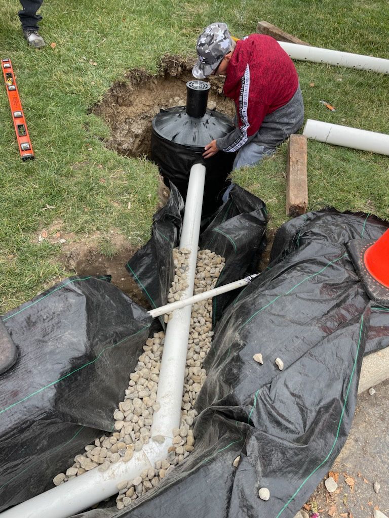 Man installing drainage system; pipes, gravel, and black fabric in a trench, on a lawn.