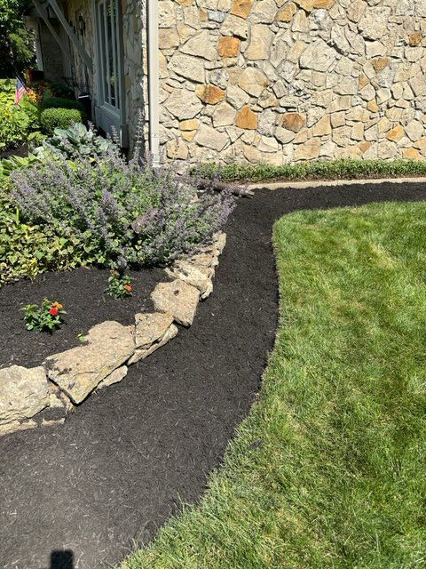 A black mulch pathway curves through green grass beside a stone wall and garden.