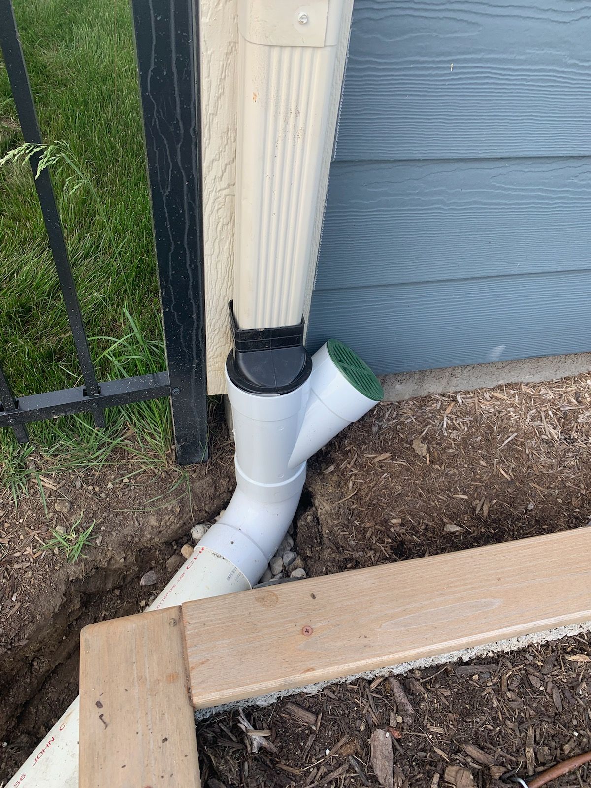 White gutter system with Y-connector draining into a buried pipe, beside a house and fence.