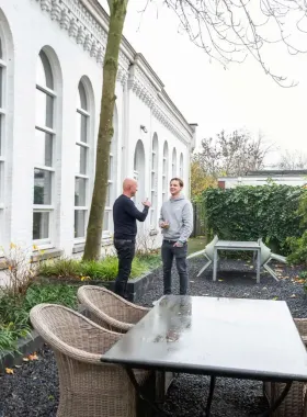 Voor een wit gebouw staan twee mannen met elkaar te praten.