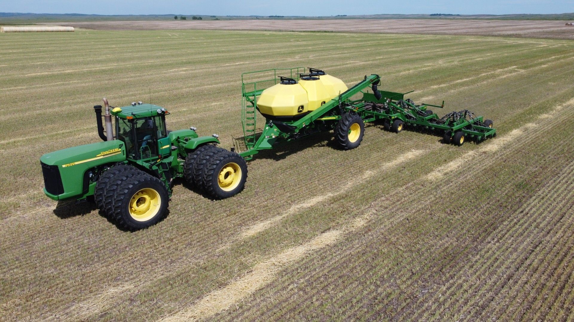 Green tractor pulling a yellow seed drill across a harvested field