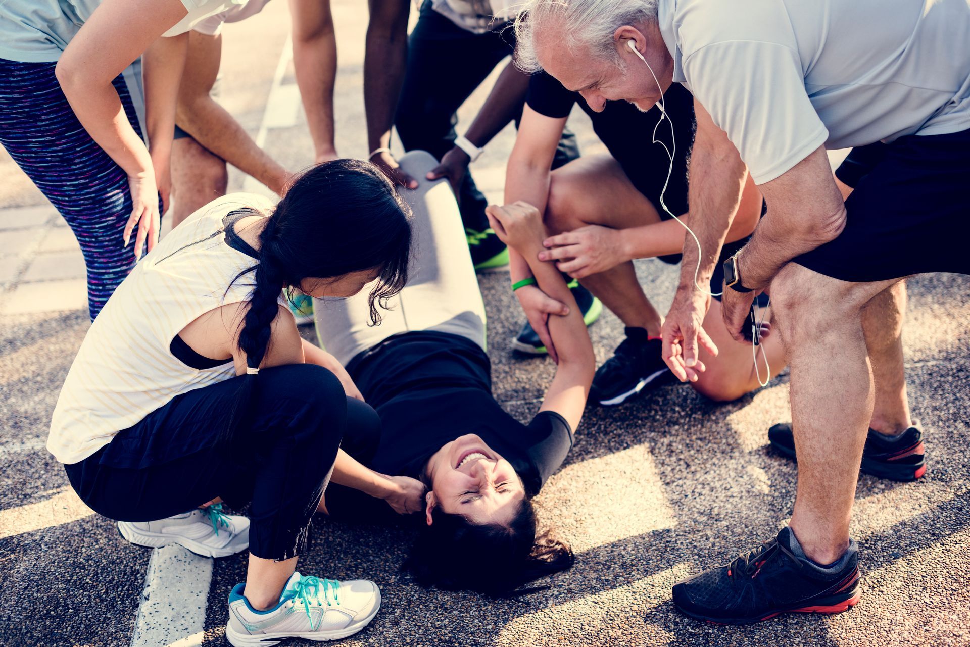 Group helping an injured woman on a paved surface; she smiles while holding her arm; others look on concerned.