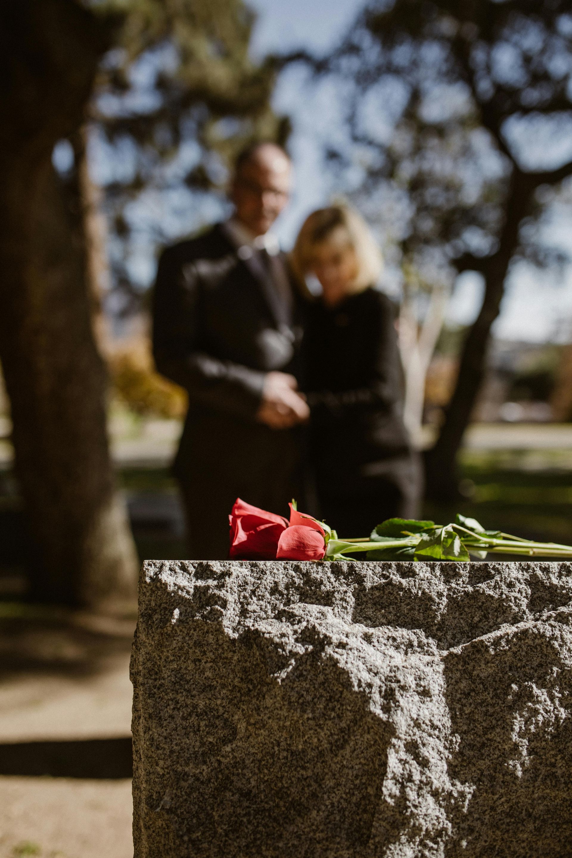 Rose on a tombstone; blurred couple in black attire stands behind in a sunny cemetery.