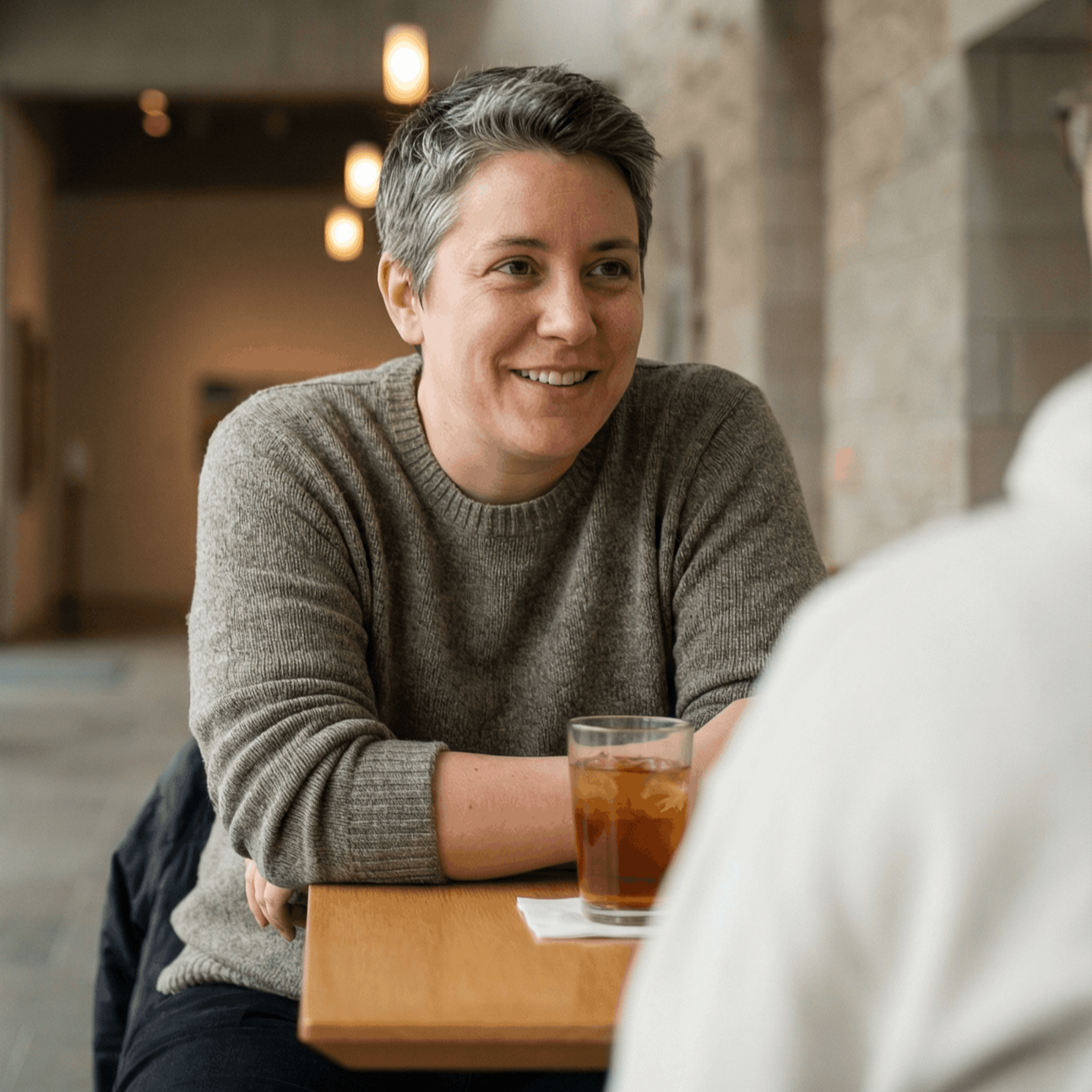 Woman at a table smiles, facing a person. A glass of iced tea sits next to her. Indoors, neutral setting.
