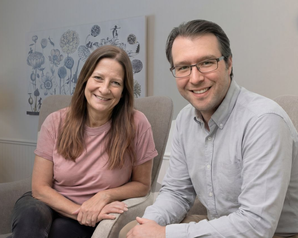 Woman and man smiling, seated indoors, near wall with floral art.