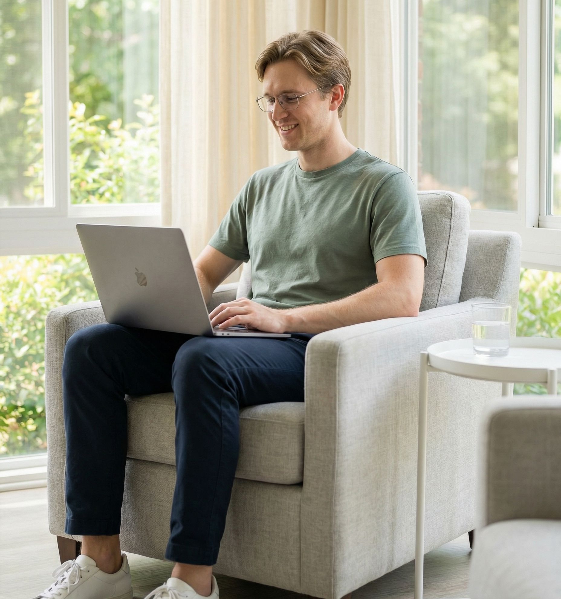 Man in glasses using laptop, seated in armchair near window. He smiles, wearing a green shirt and navy pants.