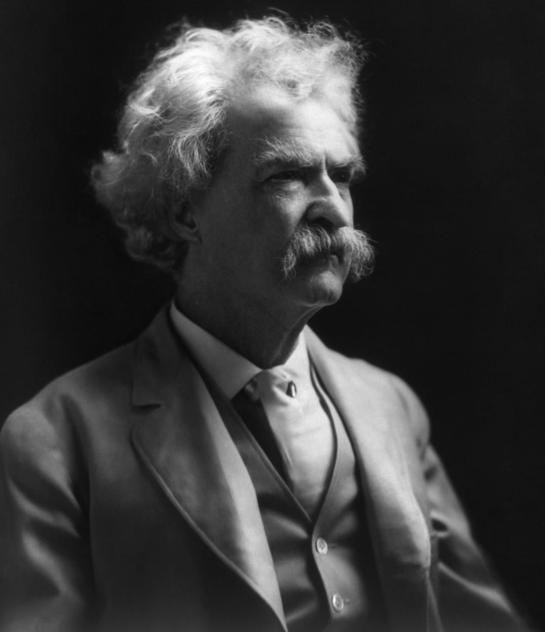 Black-and-white portrait of a man with curly white hair and a mustache in a suit, looking left.
