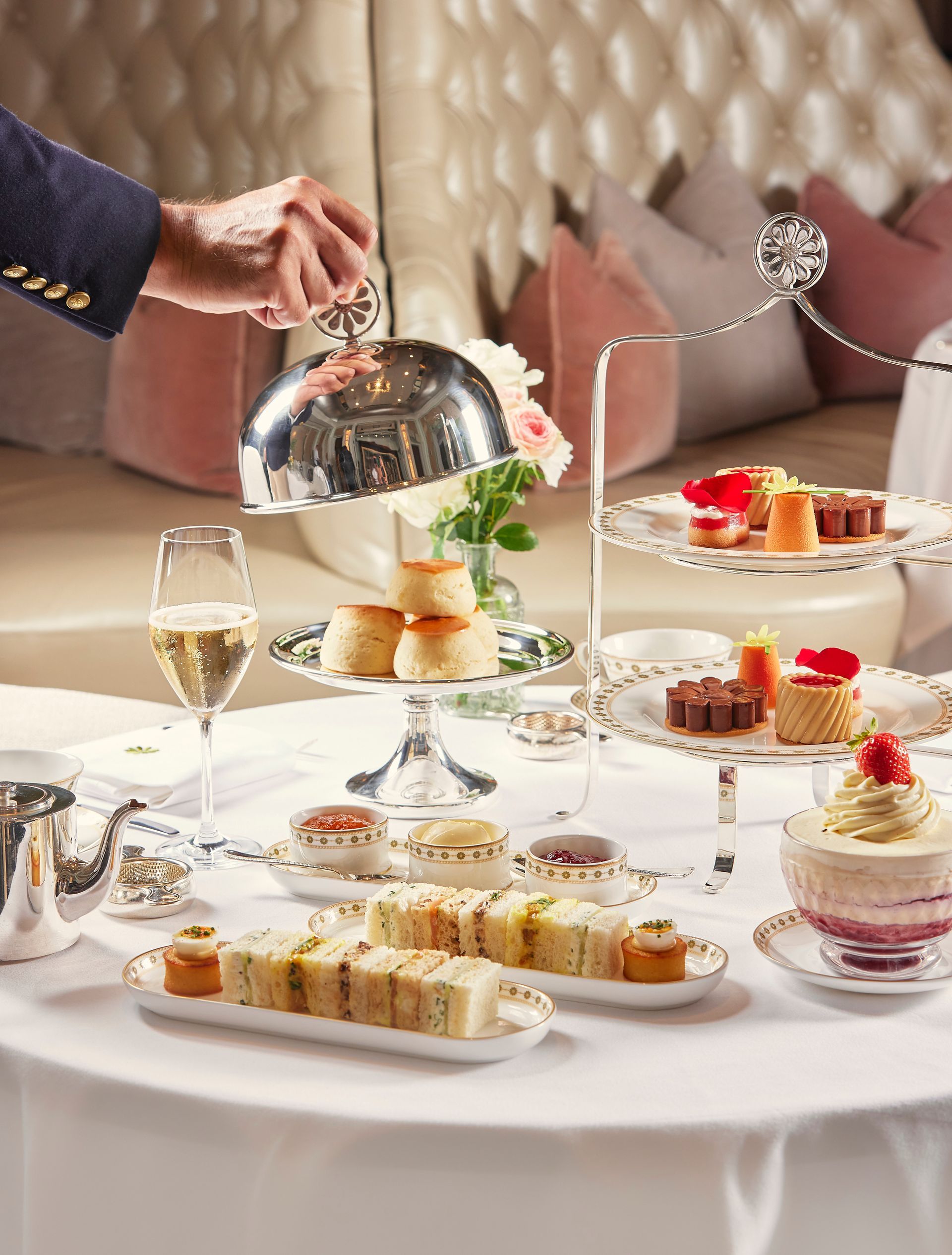 Elegant tea table with pastries, sandwiches, champagne, and a hand lifting a silver serving dome
