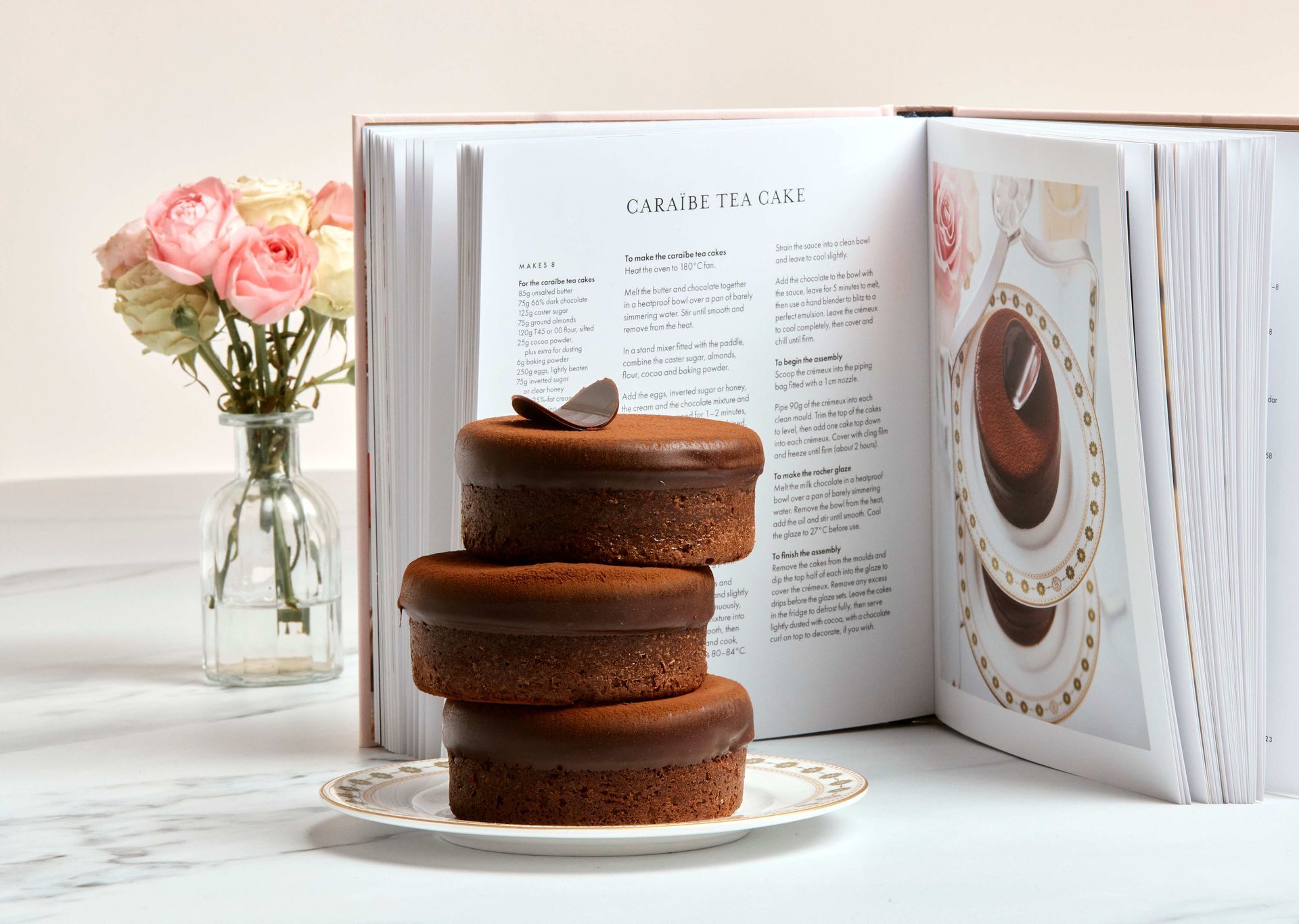 Stacked chocolate cake on a plate beside an open book and pink flowers in a bright, white setting