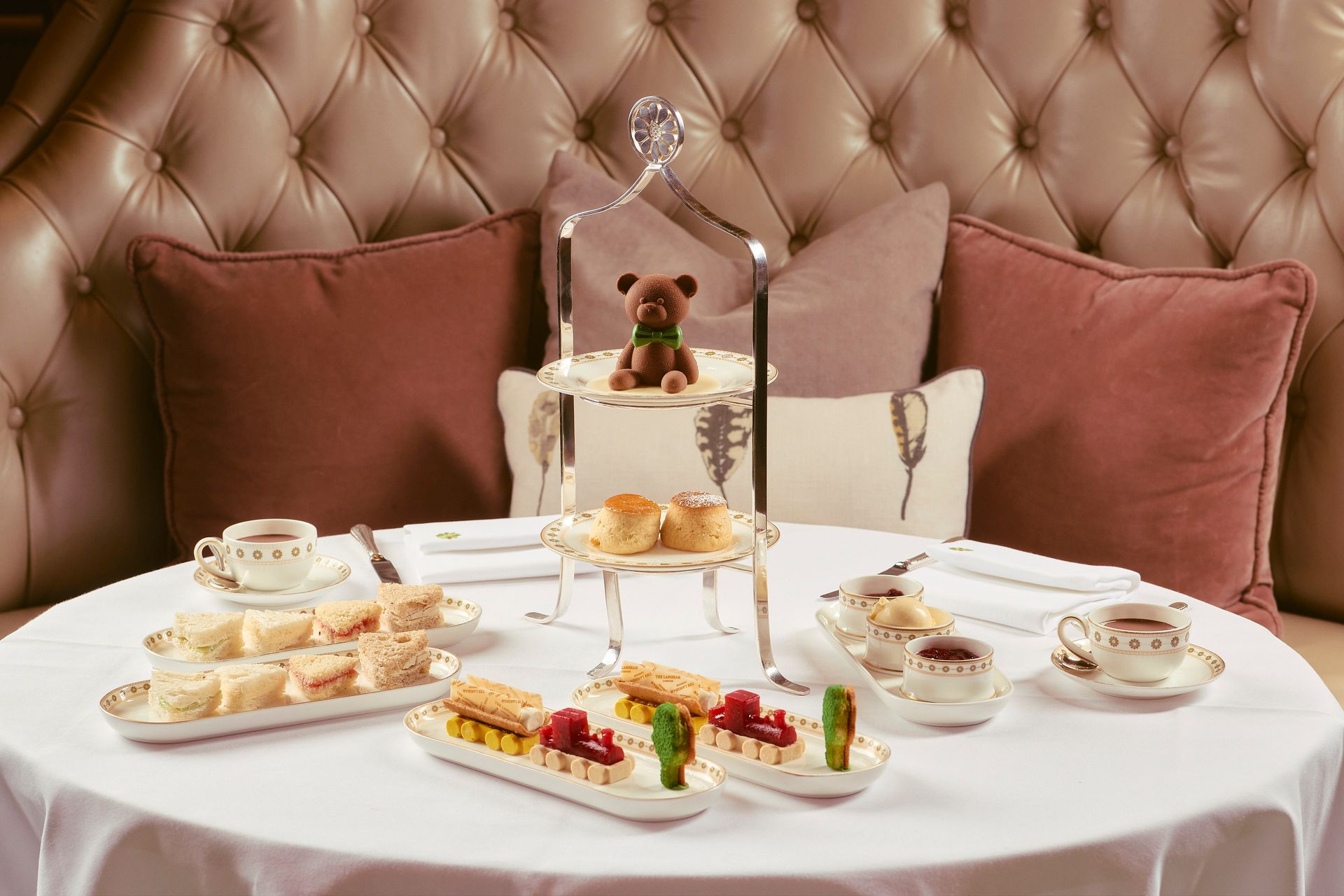 Elegant afternoon tea spread on a white table in front of a pink tufted booth with blush pillows