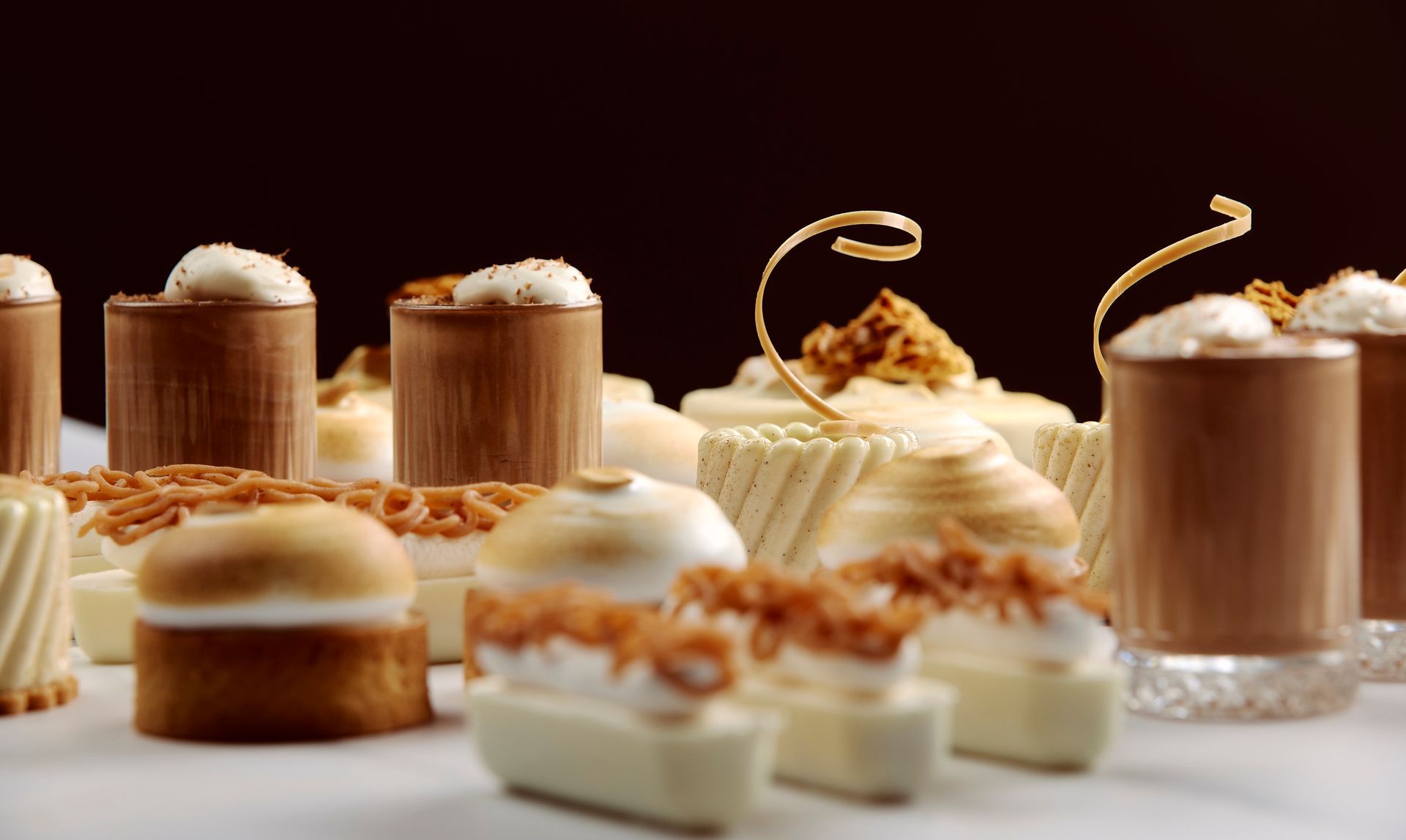 Assorted small desserts and mousse cups with whipped cream on a white table against a dark background