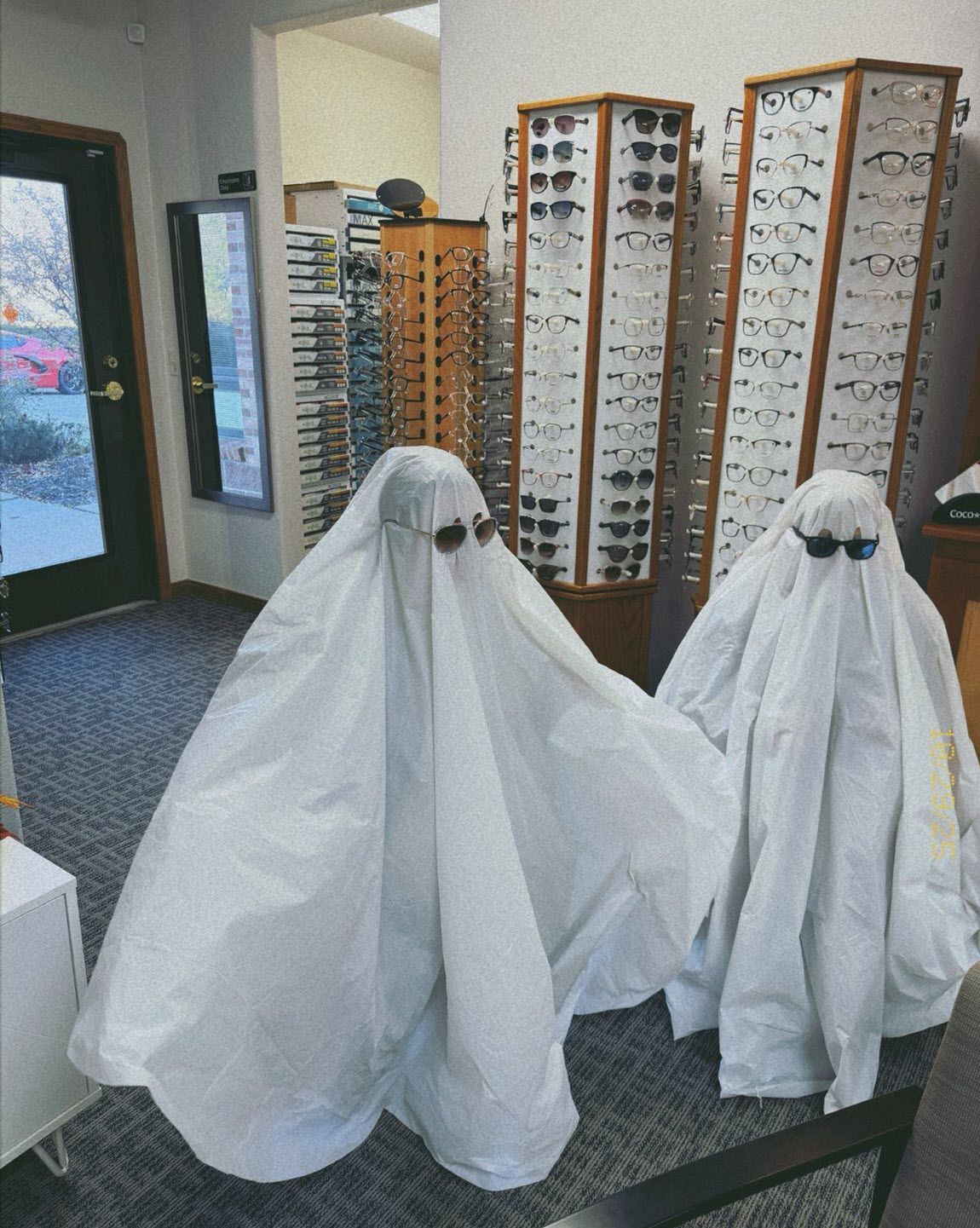 Two ghost figures wearing sunglasses in an optical shop, near displays of eyeglasses.