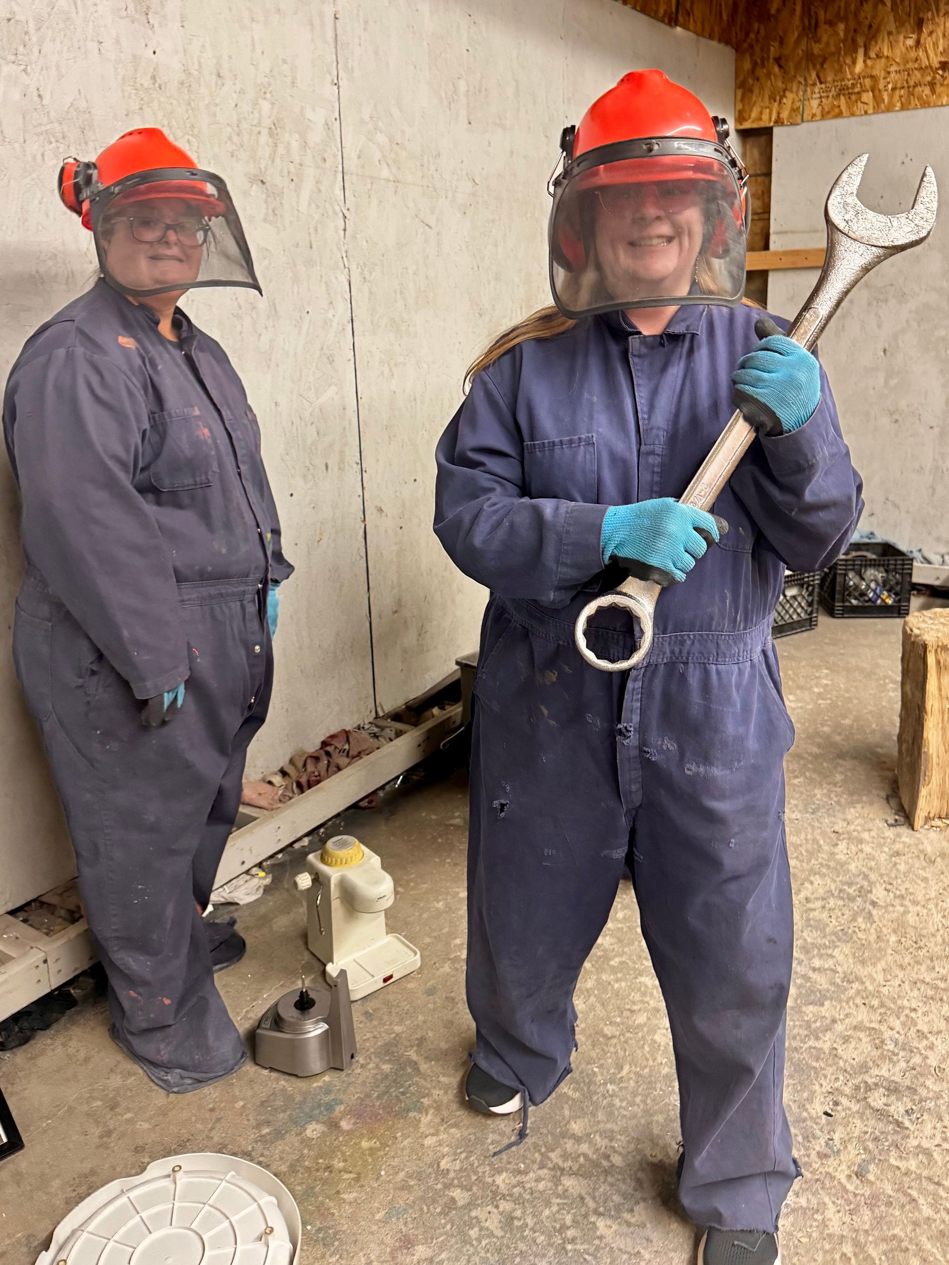 Two utility workers in red hard hats and blue coveralls, one holding a large wrench beside a wall.