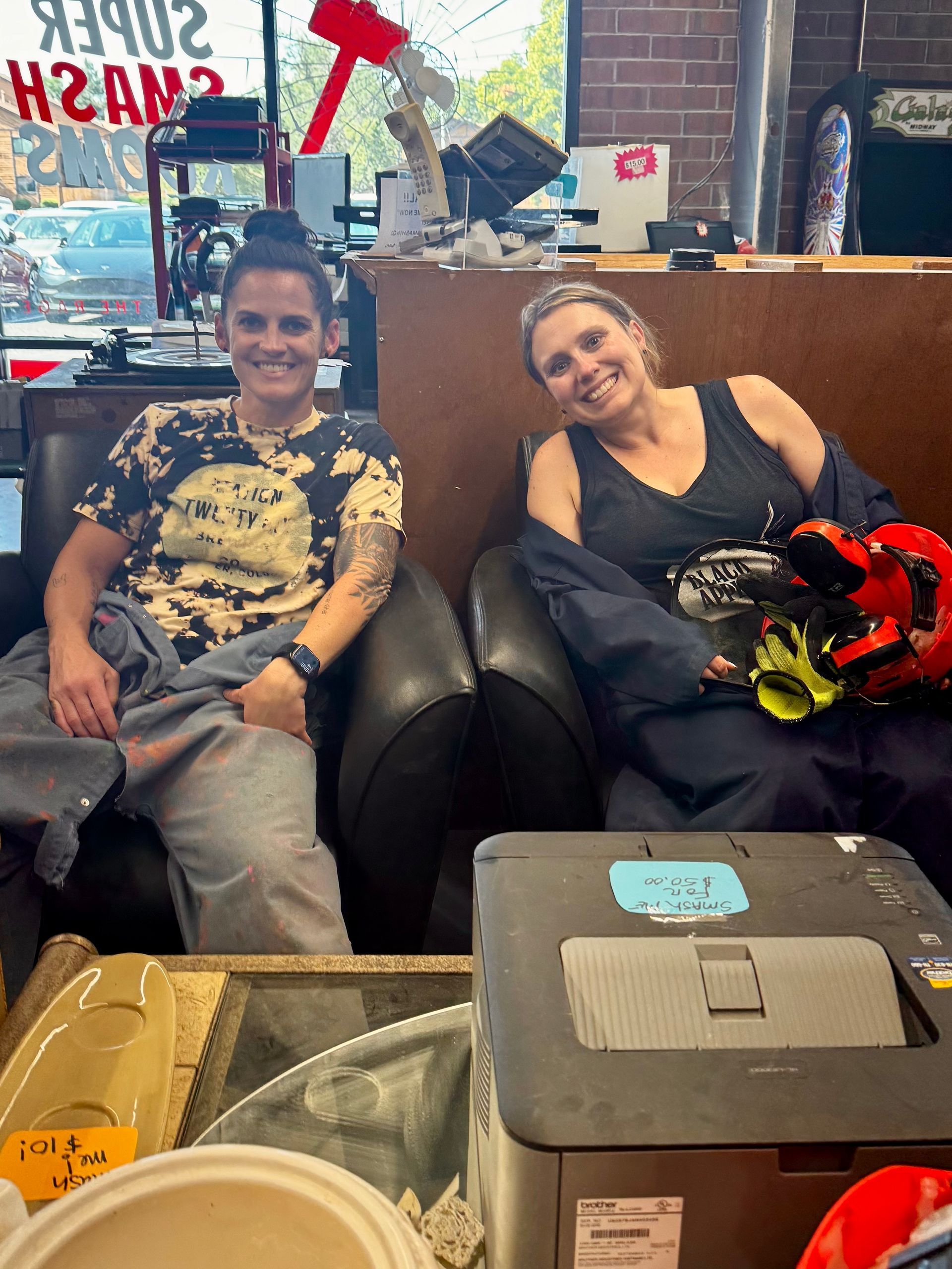 Two people smiling on black chairs in a bright room, with boxes and a red-and-black object on the table in front