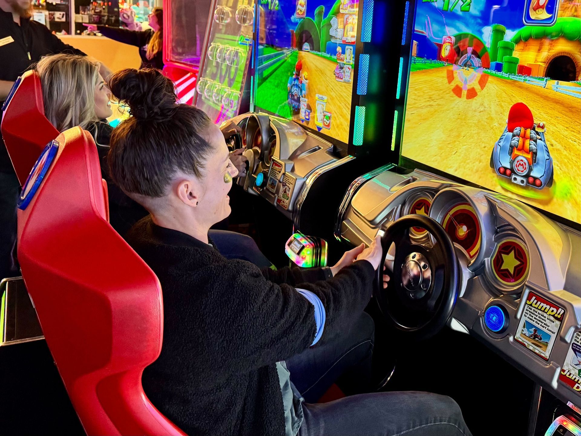 Woman playing a racing arcade game, sitting in a red seat, smiling at the screen.