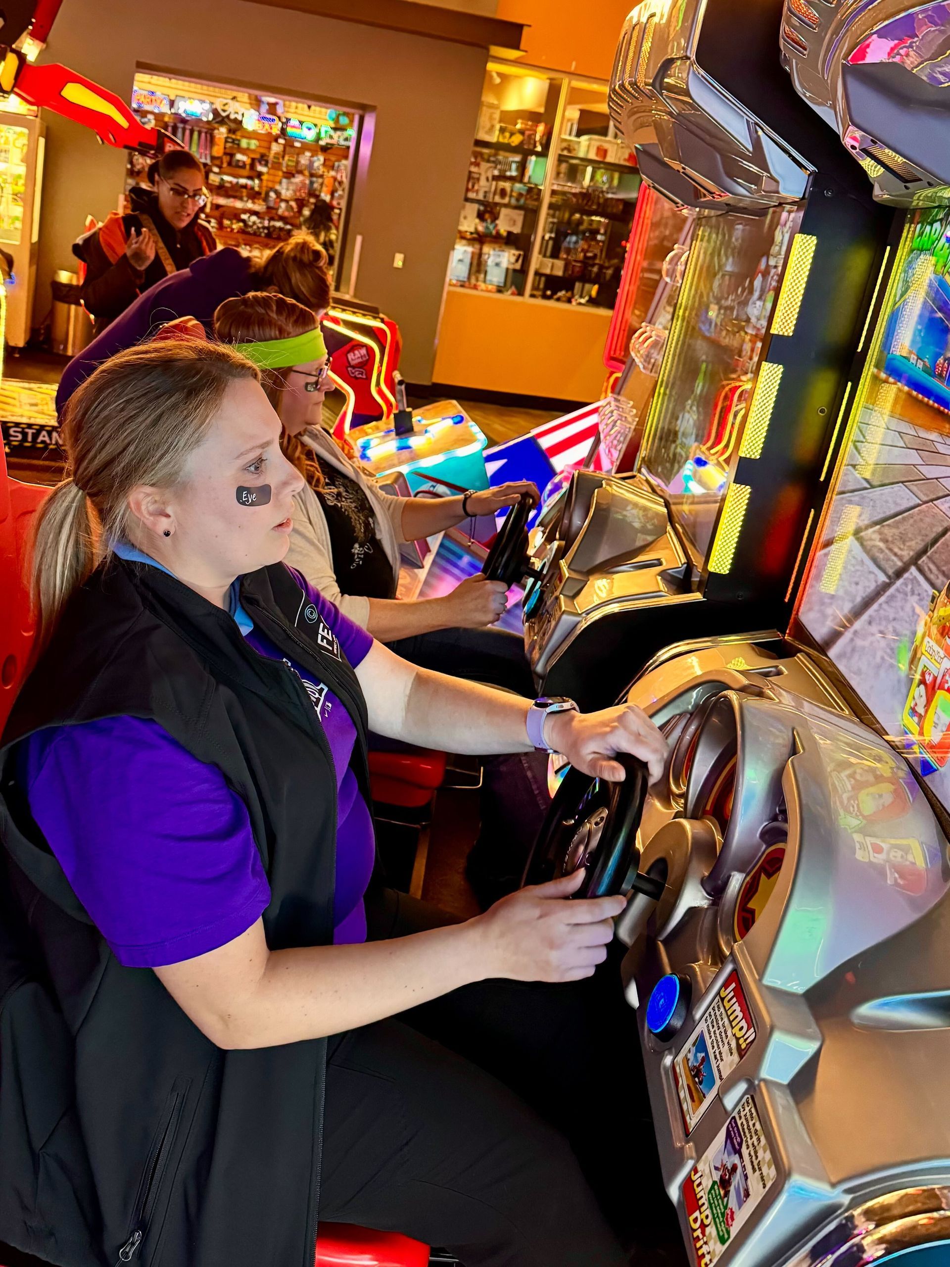 Two ladies in an arcade playing a driving game. 