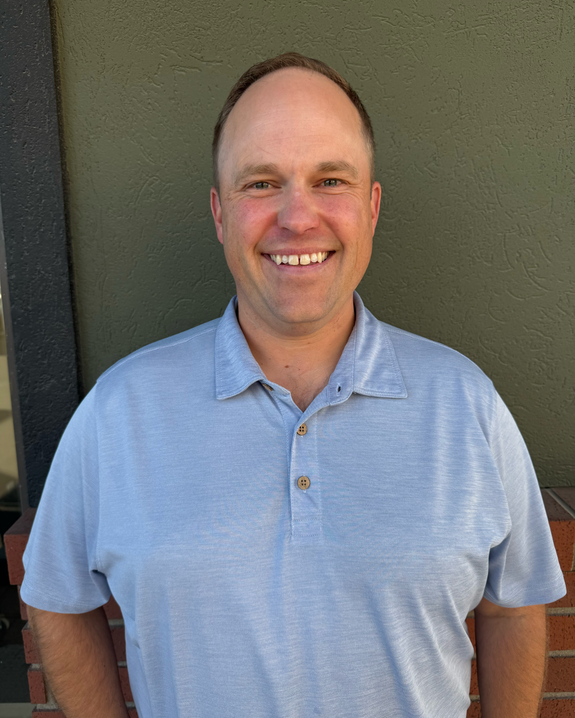 Man smiling, wearing a light blue polo shirt, standing in front of a green wall.
