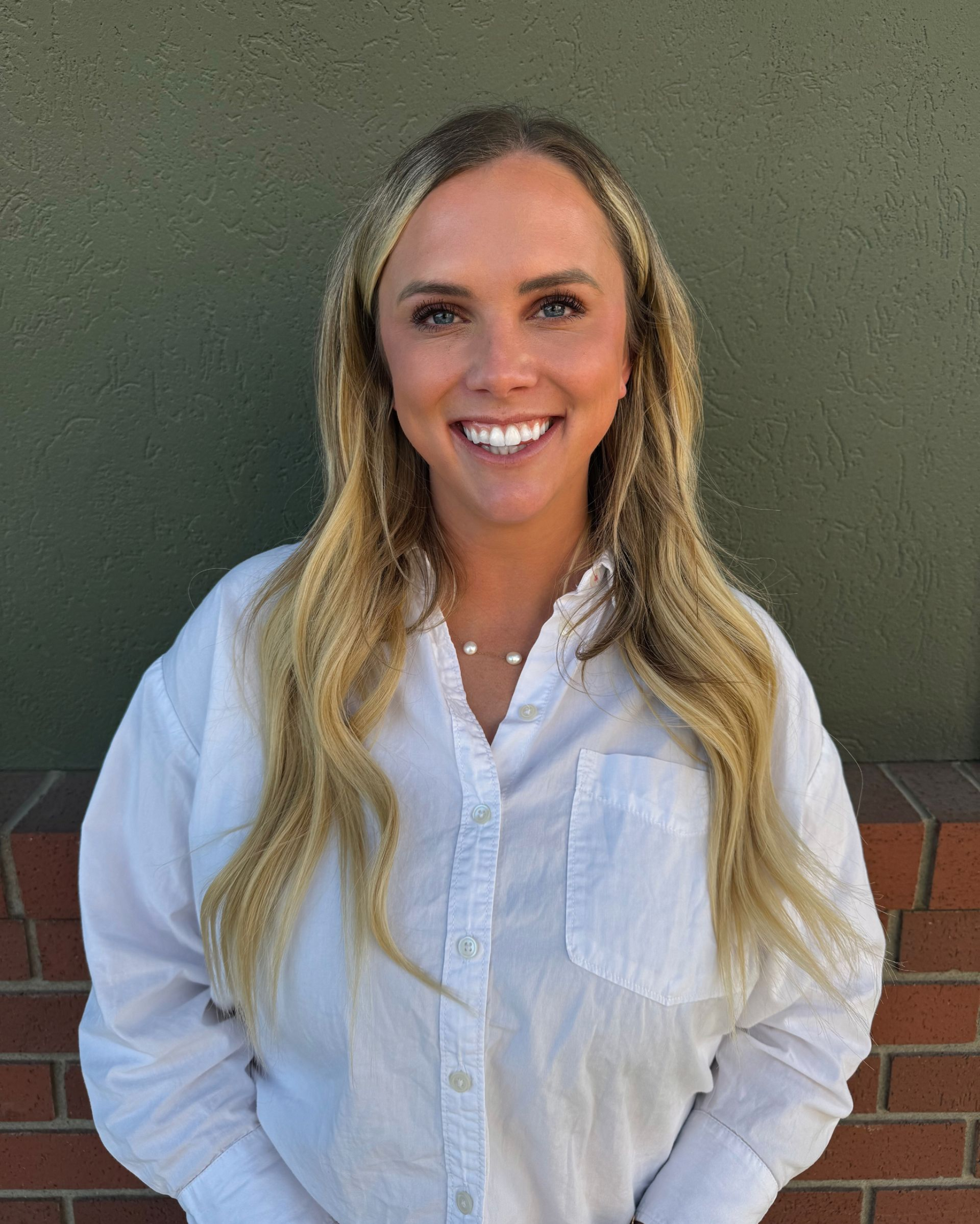 Woman with blonde hair wearing a white shirt smiles at the camera, standing in front of a green and brick wall.