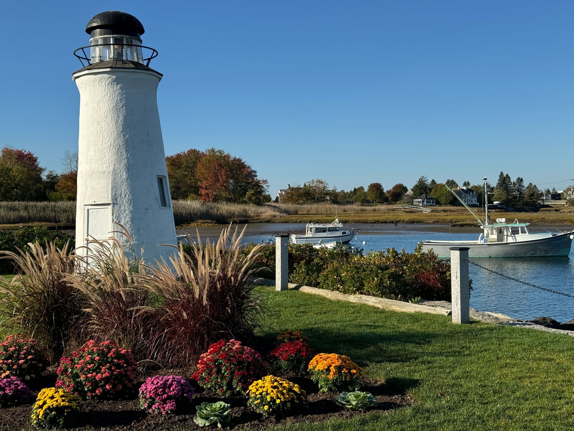 White lighthouse with black lantern on a sunny day. Boats in water, autumn foliage, and colorful flowers in foreground.